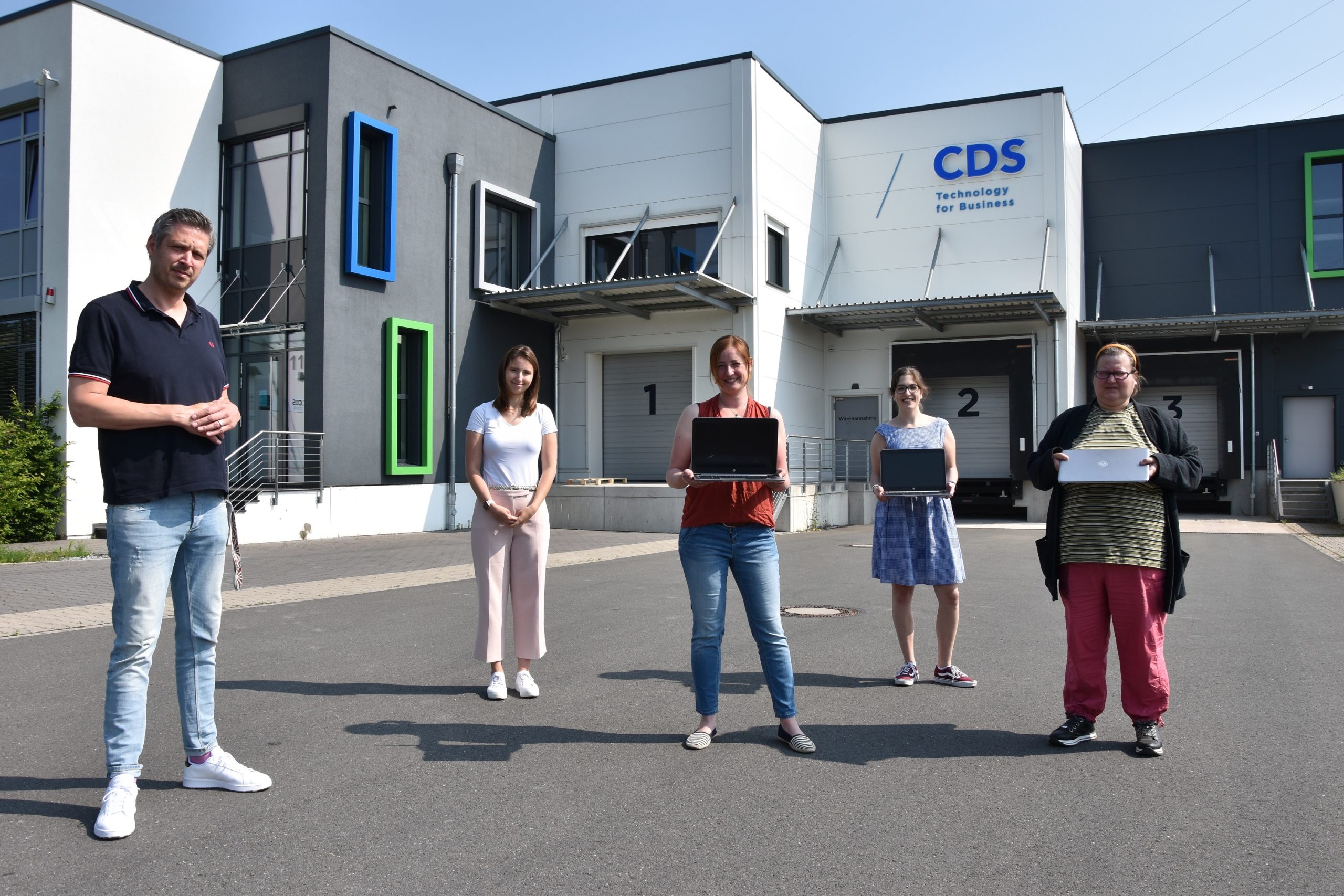 Foto, das fünf Menschen zeigt, die auf Abstand nebeneinander stehen und gespendete Laptops präsentieren